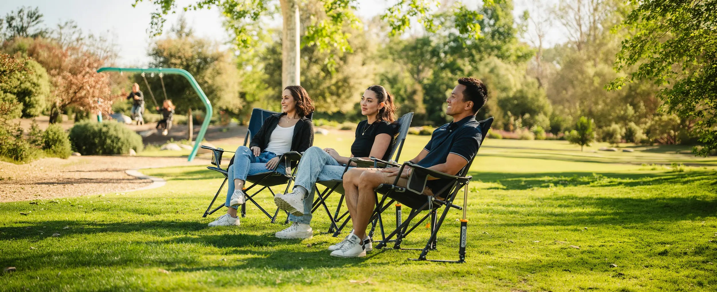 Three people sitting in Comfort Pro Rockers on a grassy park lawn, with trees and a playground visible in the background.