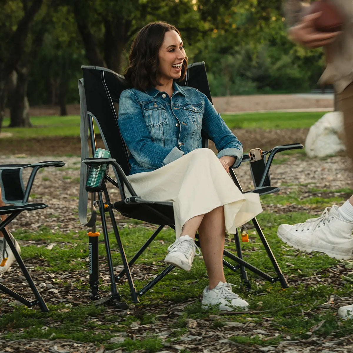 A woman sitting in a black Comfort Pro Rocker as a group of people are playing football beside her at a park. 