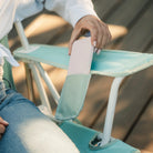 Person sliding a bottle into the side pocket of the pastel green Comfort Pro Rocker while seated outdoors.