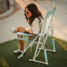 Woman relaxing in a pastel green Comfort Pro Rocker outdoors, holding a drink and seated on grass.