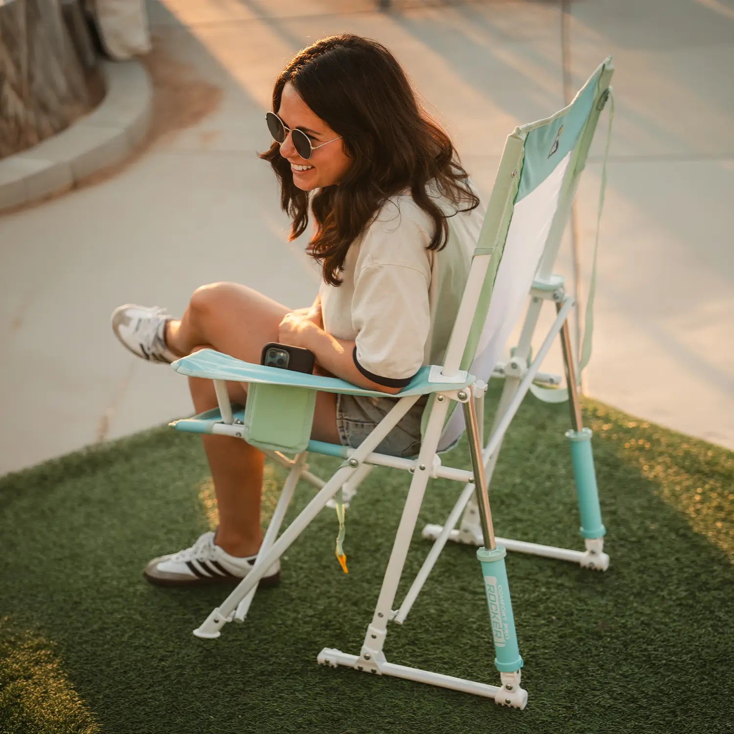 Woman relaxing in a pastel green Comfort Pro Rocker outdoors, holding a drink and seated on grass.