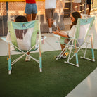 Two women carrying folded pastel green Comfort Pro Rocker chairs outdoors.