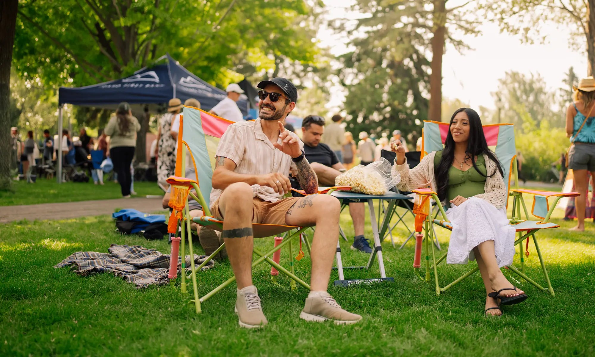 A man and a woman relax in colorful folding lawn chairs at an outdoor park picnic, surrounded by other people, trees, and a blue canopy tent in the background.