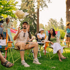 A man sitting in a sweet tea comfort pro rocker on a grassy lawn throws a bag of popcorn toward a woman in another chair at an outdoor picnic or festival. Other people and trees are visible in the background.