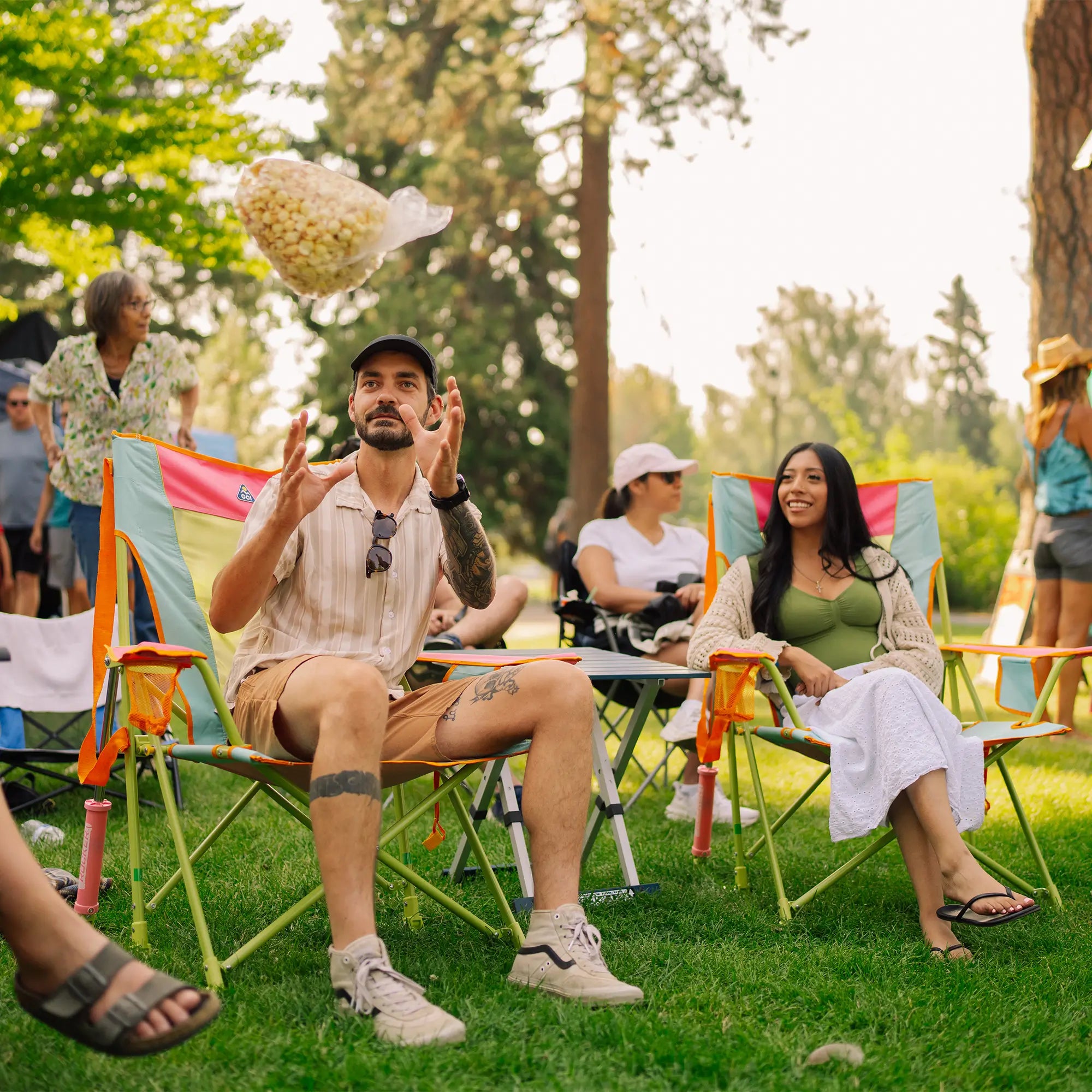 A man sitting in a sweet tea comfort pro rocker on a grassy lawn throws a bag of popcorn toward a woman in another chair at an outdoor picnic or festival. Other people and trees are visible in the background.
