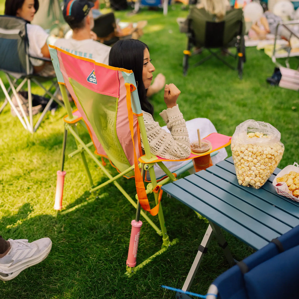 A woman sits in a sweet tea comfort pro rocker on a grassy lawn at a picnic, with a small table holding a drink and a large bag of popcorn nearby.