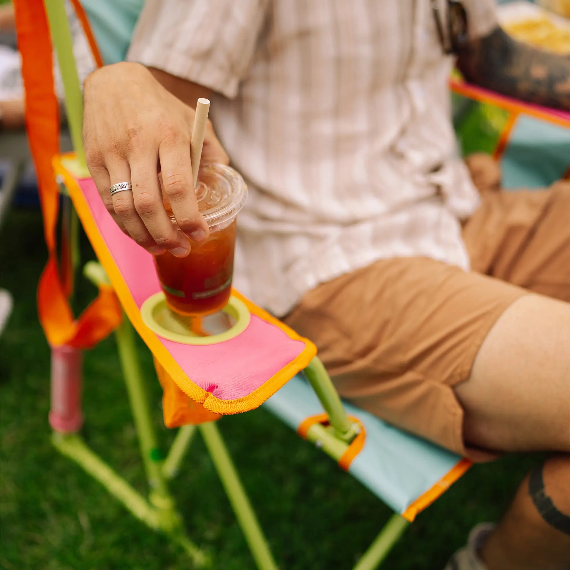 A close-up lifestyle shot of a person sitting in a sweet tea comfort pro rocker outdoors on grass, holding a plastic cup with a straw in the chair's side cup holder.