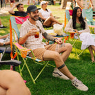 A man sitting in a sweet tea comfort pro rocker on a grassy lawn, holding an iced drink. The chair has a green frame with pink and teal fabric panels and orange side straps.