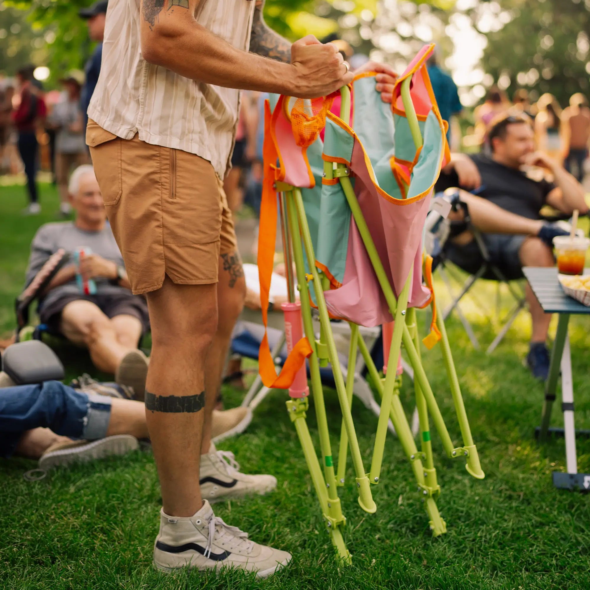 A person outdoors on grass holding a sweet tea comfort pro rocker. Other people and a picnic-style outdoor setting are visible in the background.