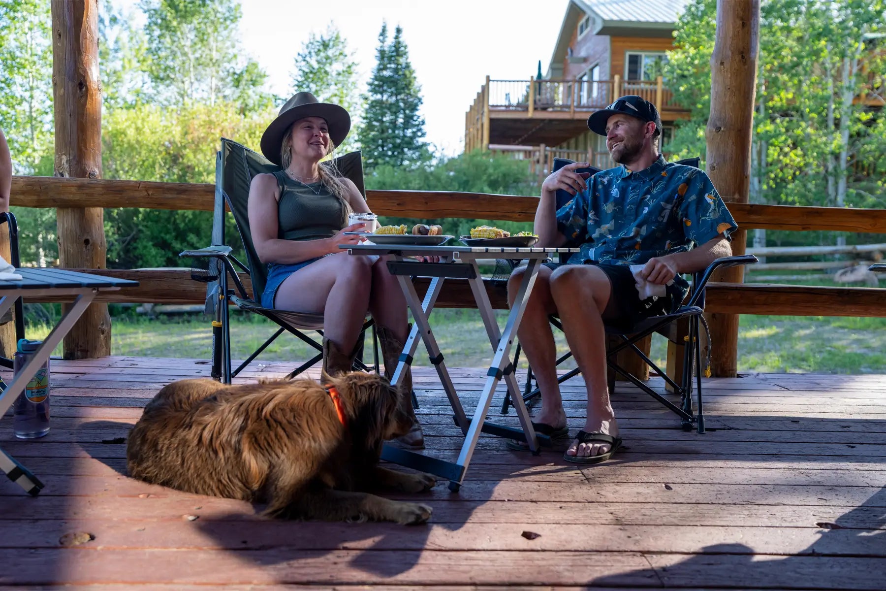 A couple relaxes on a porch, sitting in Comfort Pro Rockers while enjoying food and conversation. A brown dog rests nearby, adding to the cozy vibe.