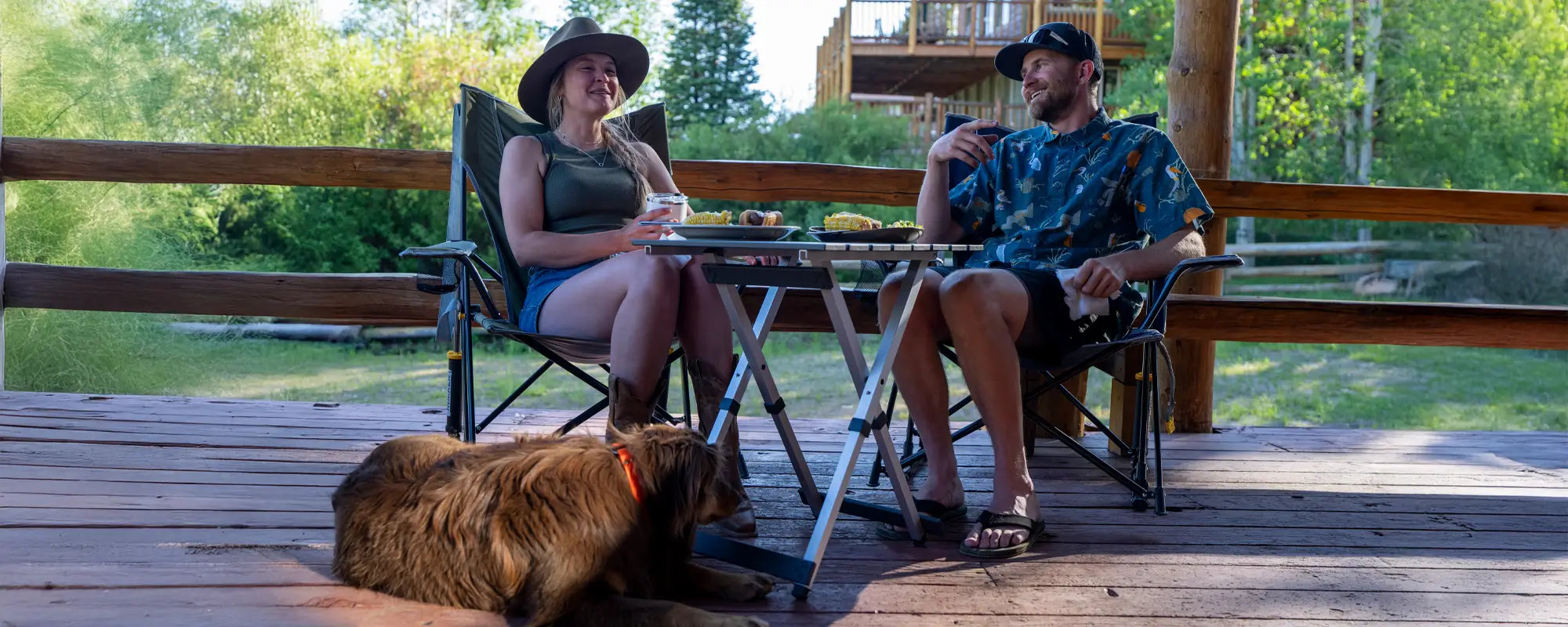 A couple relaxes on a porch, sitting in Comfort Pro Rockers while enjoying food and conversation. A brown dog rests nearby, adding to the cozy vibe.