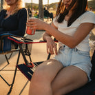 A woman placing her soda in the cupholder of the indigo/red Comfort Quad Chair. 