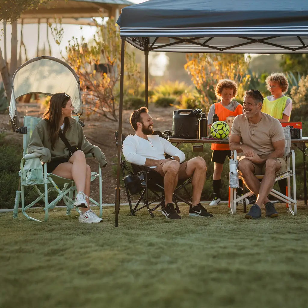 A group of parents relax under a LevrUp Canopy, seated in comfortable outdoor chairs, while kids play soccer nearby. The setup includes a SunShade Rocker, Freestyle Rocker, and a Compact Camp Chair, offering shade and comfort for game-day spectators.