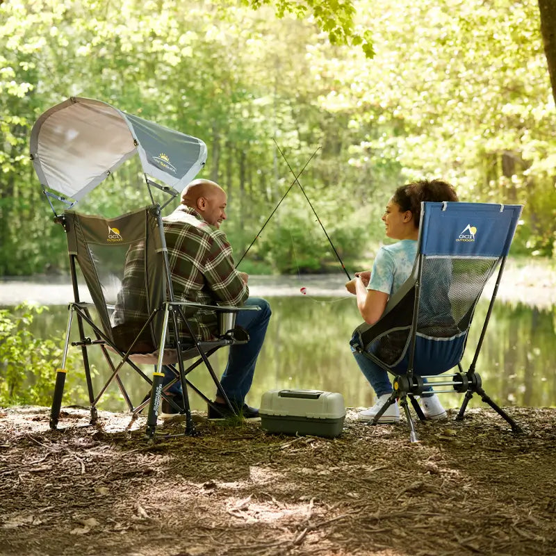 Two people fishing lakeside while seated in ComPack Rocker chairs, one with a sunshade canopy and the other with a mesh backrest.