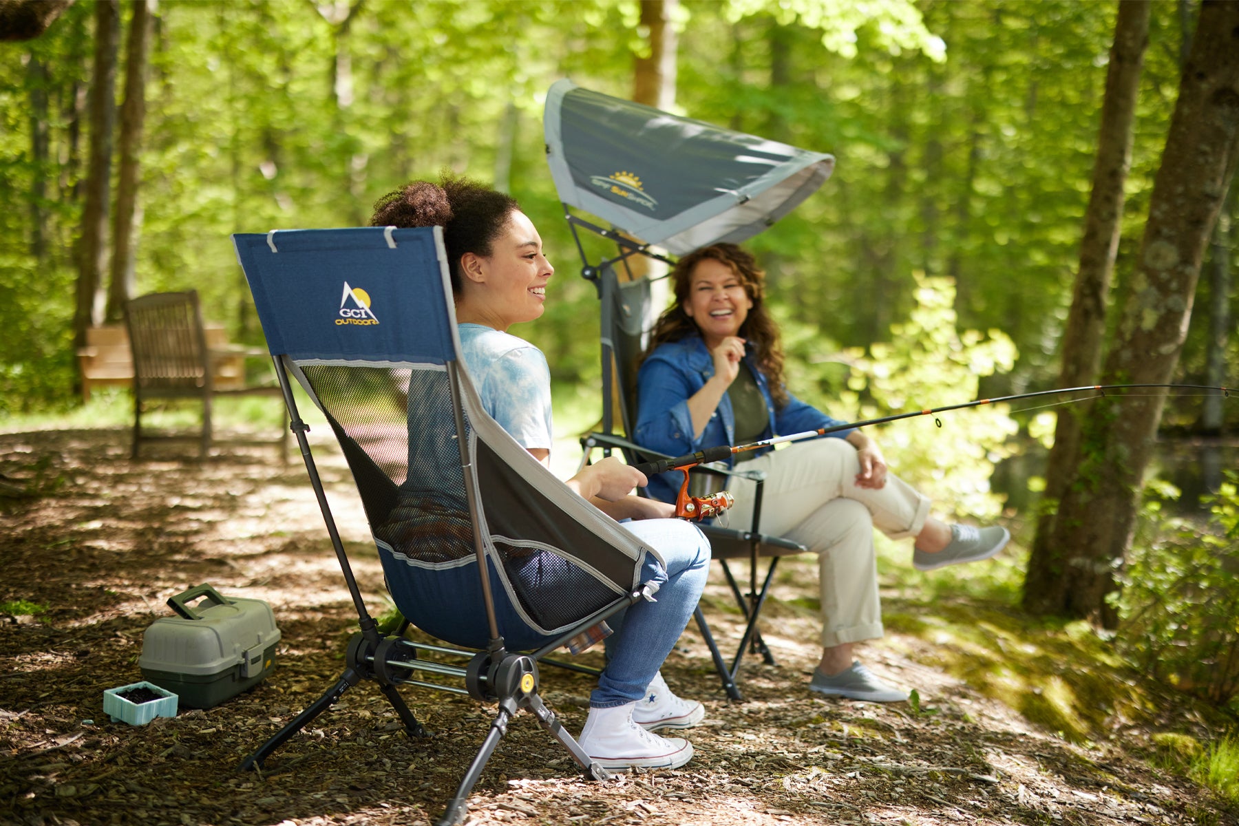 Two women sit in rocking chairs in a forest, one holding a fishing rod, showing the Compack Rocker's convenience and portability.