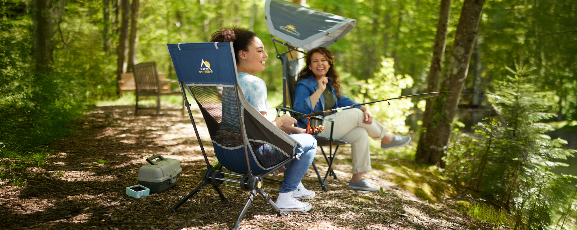 Two women sit in rocking chairs in a forest, one holding a fishing rod, showing the Compack Rocker's convenience and portability.