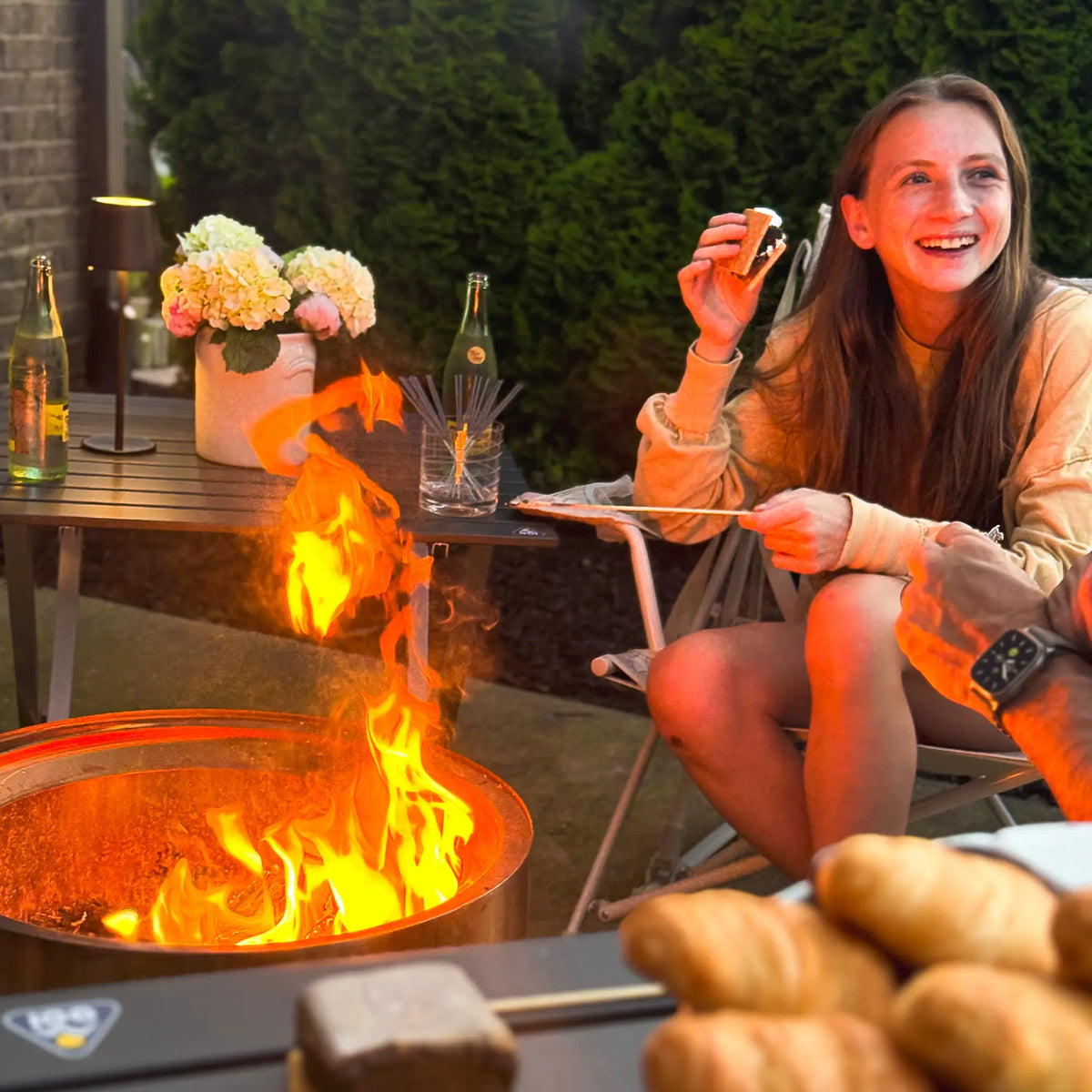 Woman sitting by a fire pit with drinks and food, smiling outdoors.
