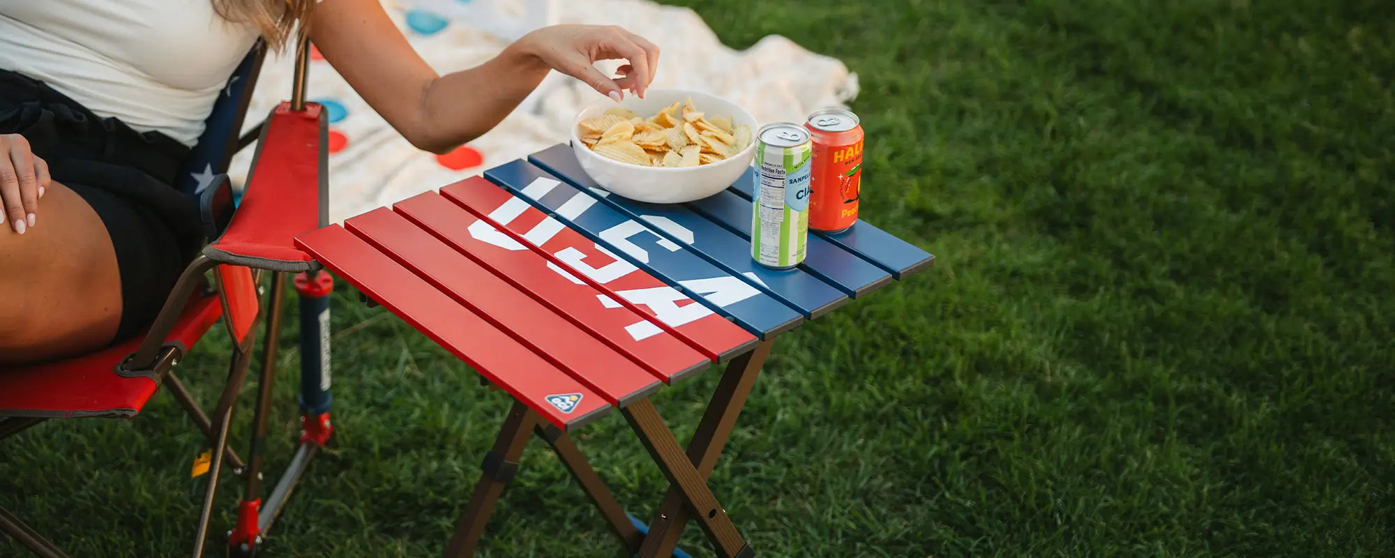 A woman reaching for a bowl of chips that are sitting on a USA Compact Camp Table. 