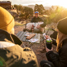 A group of friends sits around a Compact Camp Table in the desert at sunset, playing cards and sipping drinks, while one person holds a fluffy dog on their lap.