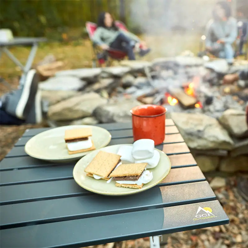 A close-up of the Compact Camp Table beside a fire pit, topped with graham crackers, toasted marshmallows, and a red mug, while people sit around the fire in the background.