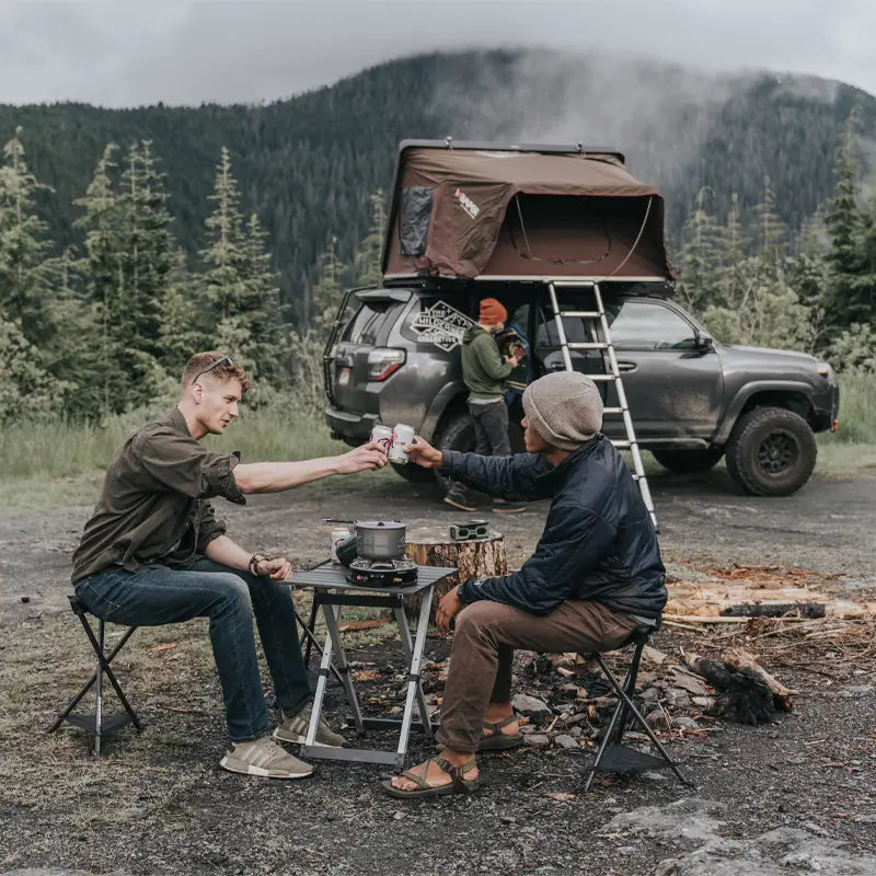 wo men sit on low stools at a campsite in front of a roof-top tent, clinking drinks over a Compact Camp Table that holds a pot on a camp stove and a few essentials.

