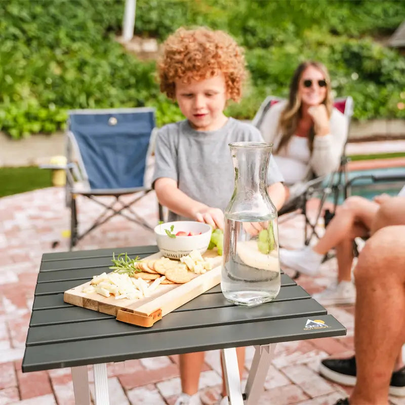 A Compact Camp Table in the foreground holds a wooden snack tray, a bowl of dip, and a glass carafe of water, while a child and adults relax poolside in the background.