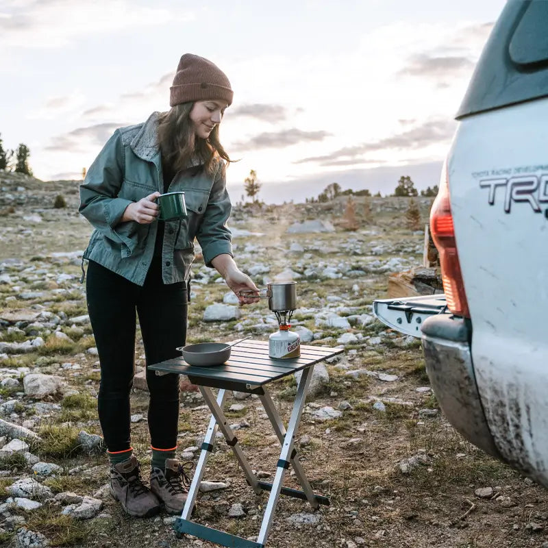 A woman at a rocky campsite brews coffee on a Compact Camp Table using a camp stove, holding a green mug in one hand with a truck tailgate nearby.