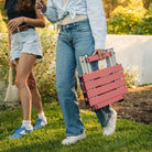 Two women walking in a backyard setting while one woman carries a coral Compact Camp Table. 