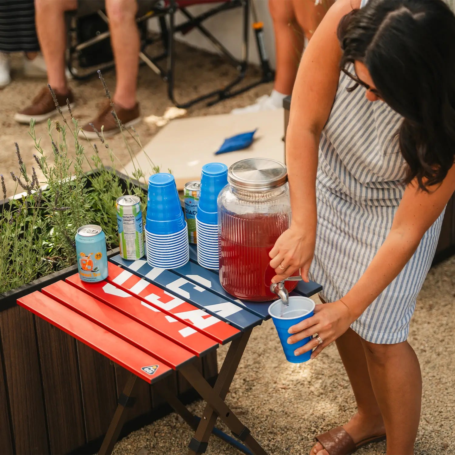 A woman pouring juice into a cup that is positioned on top of the USA Deluxe Compact Camp Table. 
