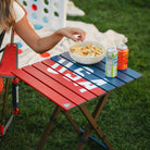 A woman reaching for a bowl of snacks that are resting on top of the USA Deluxe Compact Camp Table. 
