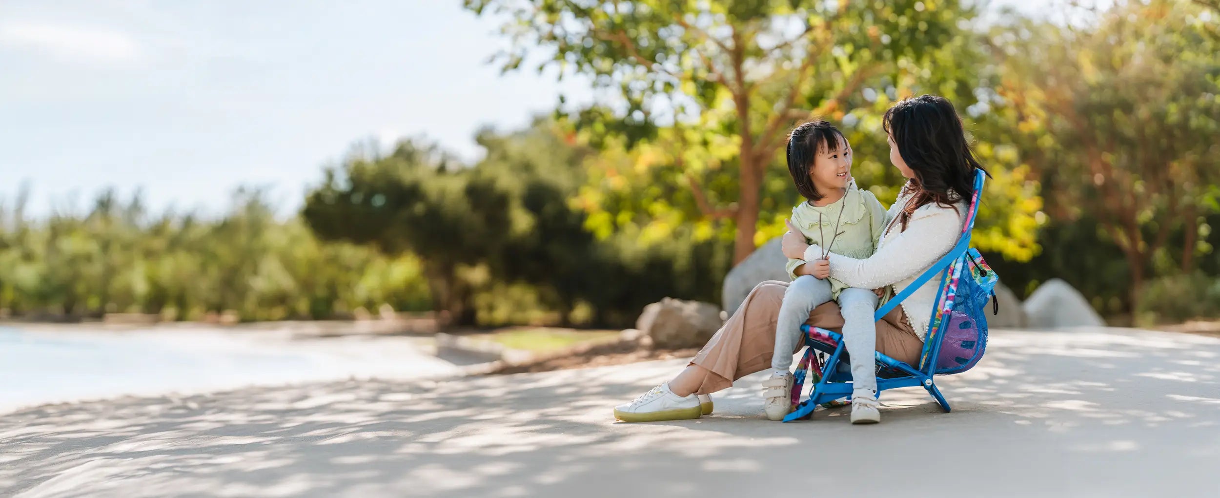 A woman holding a child while sitting in the tie dye Everywhere Chair 2 in a park. 