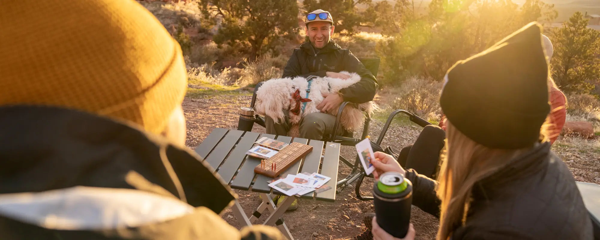 Friends play cards at sunset around a compact camp table, with drinks and a dog, highlighting the table’s practical size.