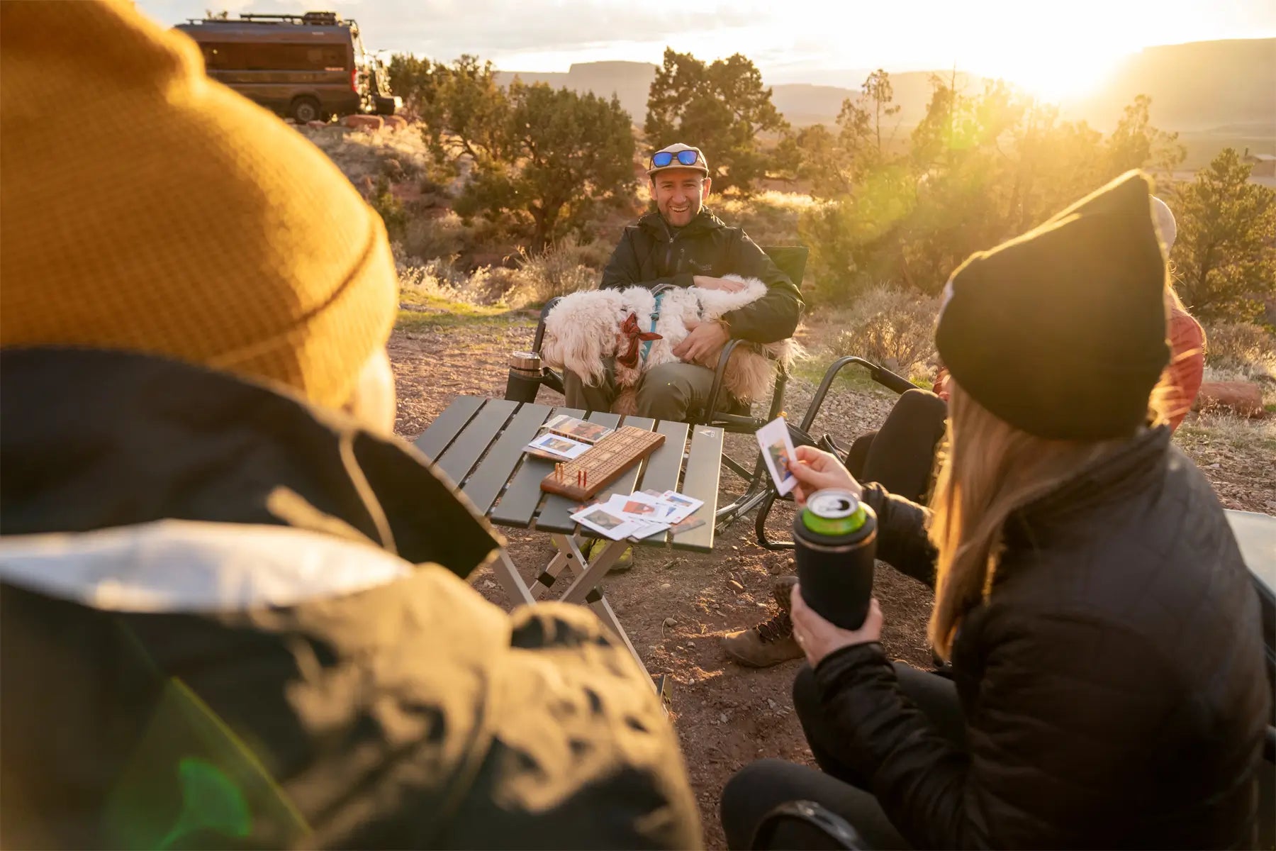 Friends play cards at sunset around a compact camp table, with drinks and a dog, highlighting the table’s practical size.