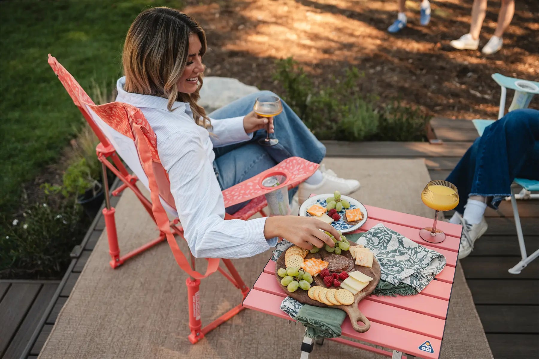 A woman reaching for a charcuterie tray sitting on a coral Compact Camp Table while she rocks in a coral Comfort Pro Rocker. 
