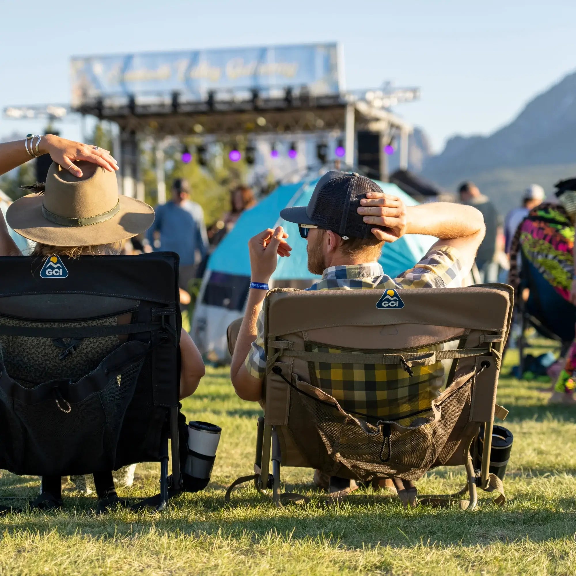 Two people relax in Everywhere Chair 2 seats at an outdoor festival, enjoying the live music. The portable chairs feature breathable mesh backs, backpack straps, and convenient storage pockets for drinks and essentials.