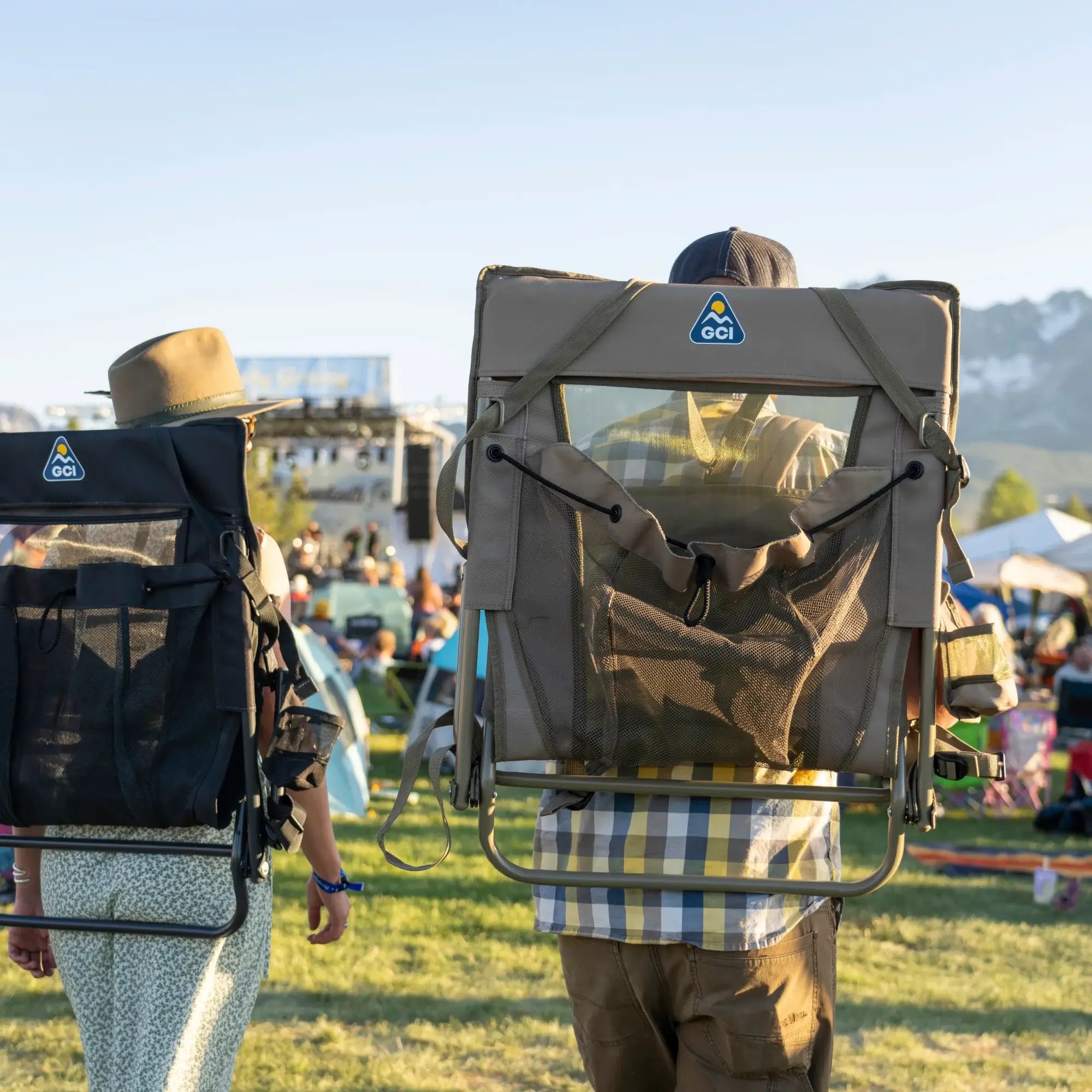 Two people walk through a festival carrying the Everywhere Chair 2 on their backs using its built-in backpack straps. The lightweight chair features a breathable mesh panel and storage pocket, making it ideal for outdoor events.