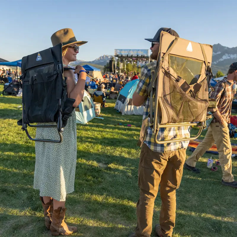 Two people walk through a festival carrying the Everywhere Chair 2 on their backs using its built-in backpack straps. The lightweight, portable design makes it easy to transport for outdoor events.