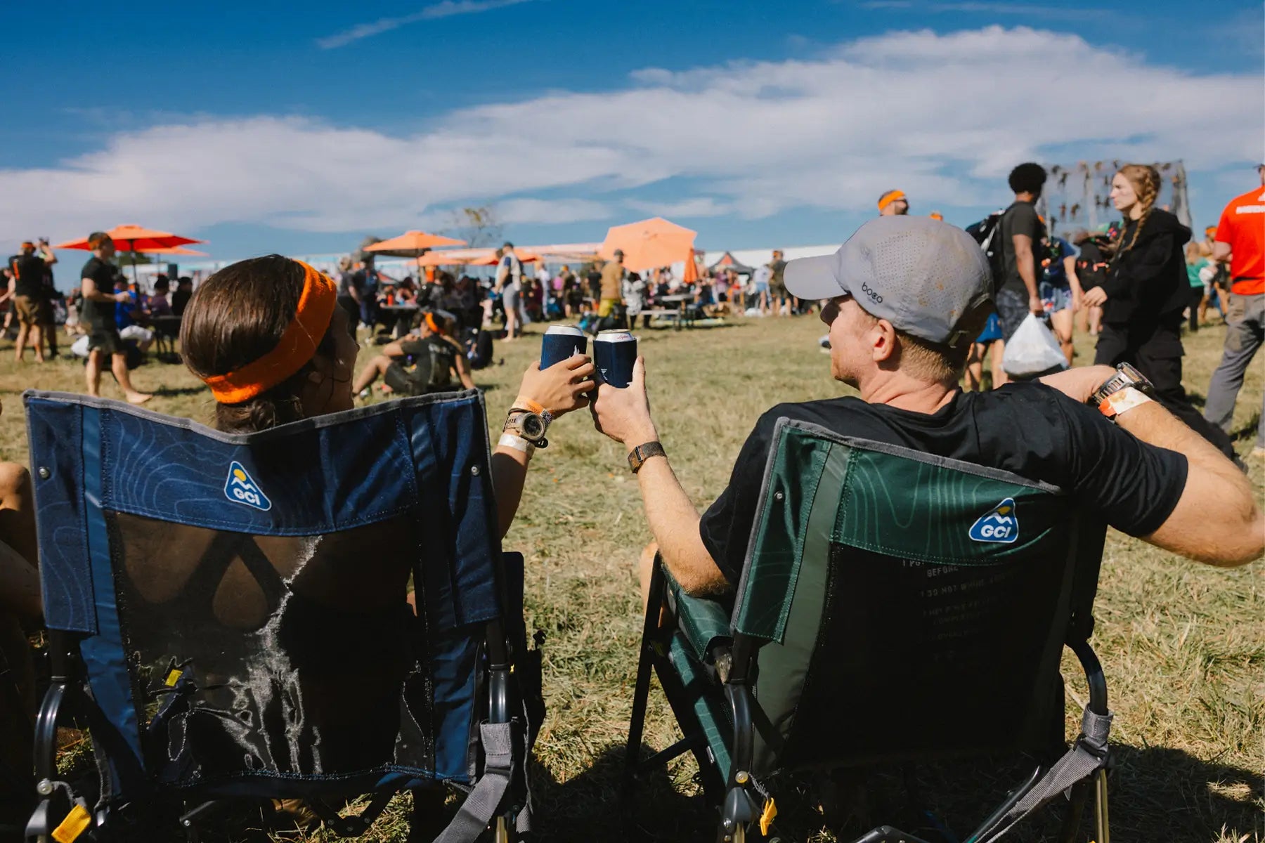 Two people sitting in Stowaway Rockers at an outdoor festival, raising drinks, with tents and a crowd in the background.