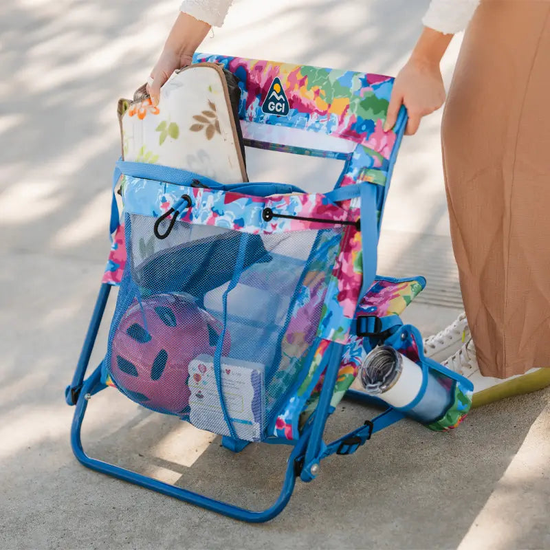 Person lifting a tie dye Everywhere Chair 2 filled with beach gear, including a helmet, towel, and seat cushion.