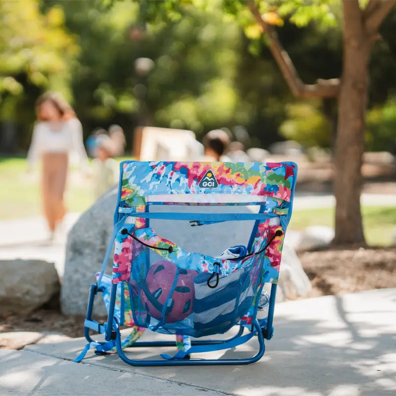 Tie dye Everywhere Chair 2 set on a shaded path, loaded with a pink helmet, towel, and other essentials in the mesh back pocket.