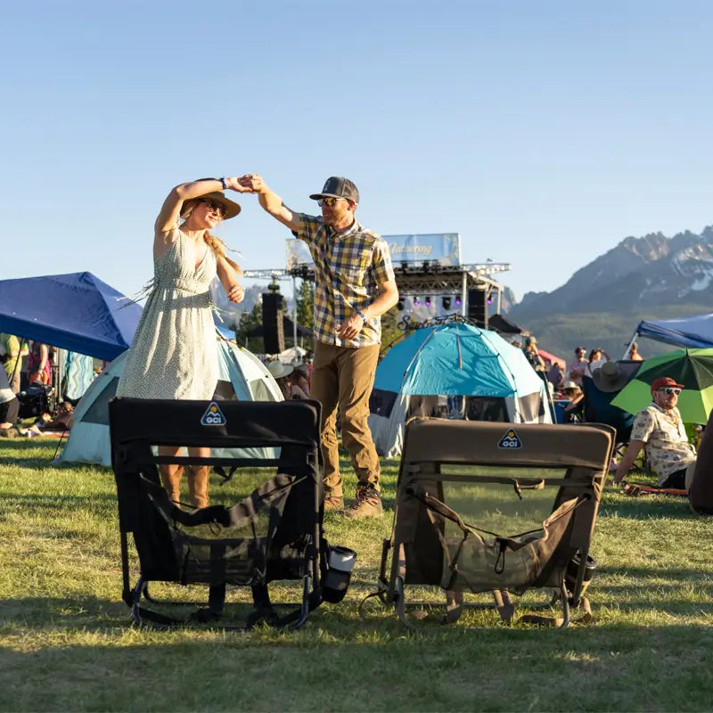 Two people dancing at an outdoor festival with black and aged fatigue Everywhere Chair 2 seats in the foreground on grassy ground.