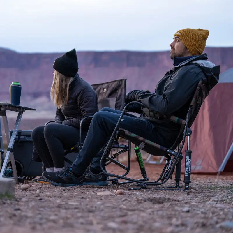Man lounging in a Firepit Rocker near a tent, gazing out across a canyon landscape while holding a drink.