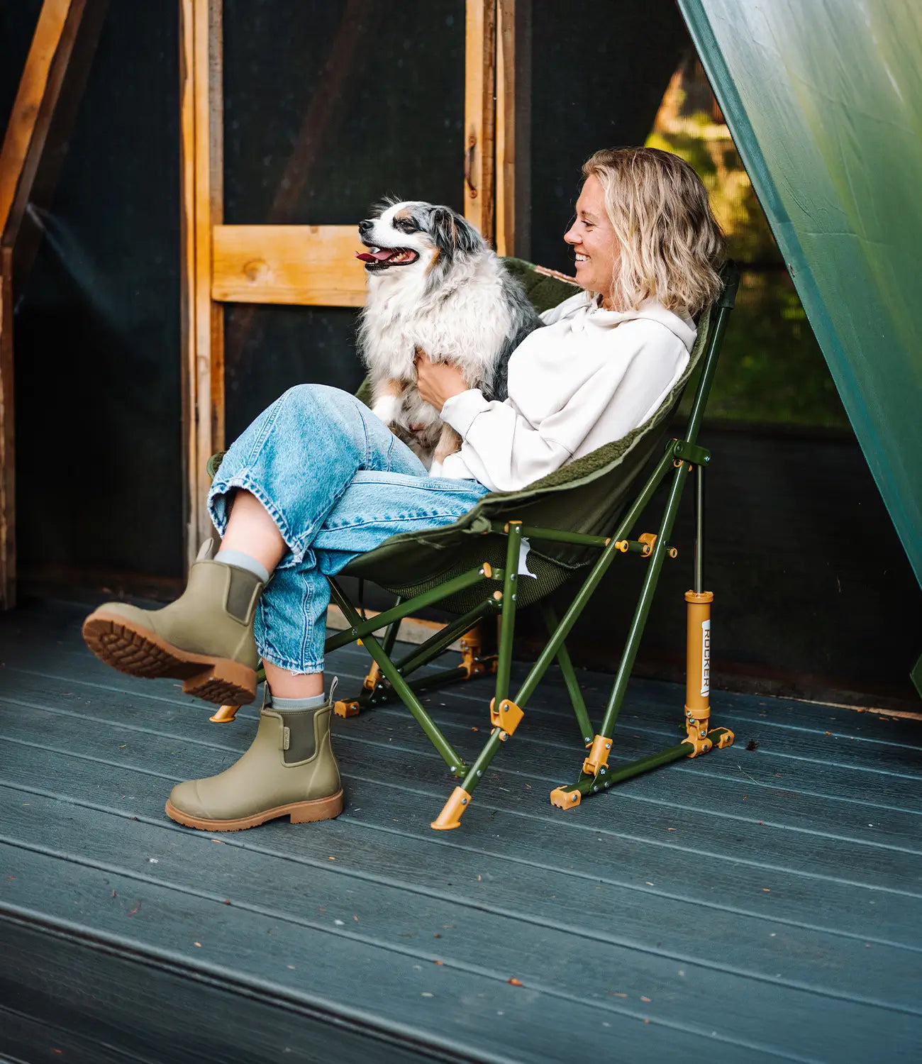 A woman holding her dog while sitting on an olive Fleece Rocker chair next to a cabin. 