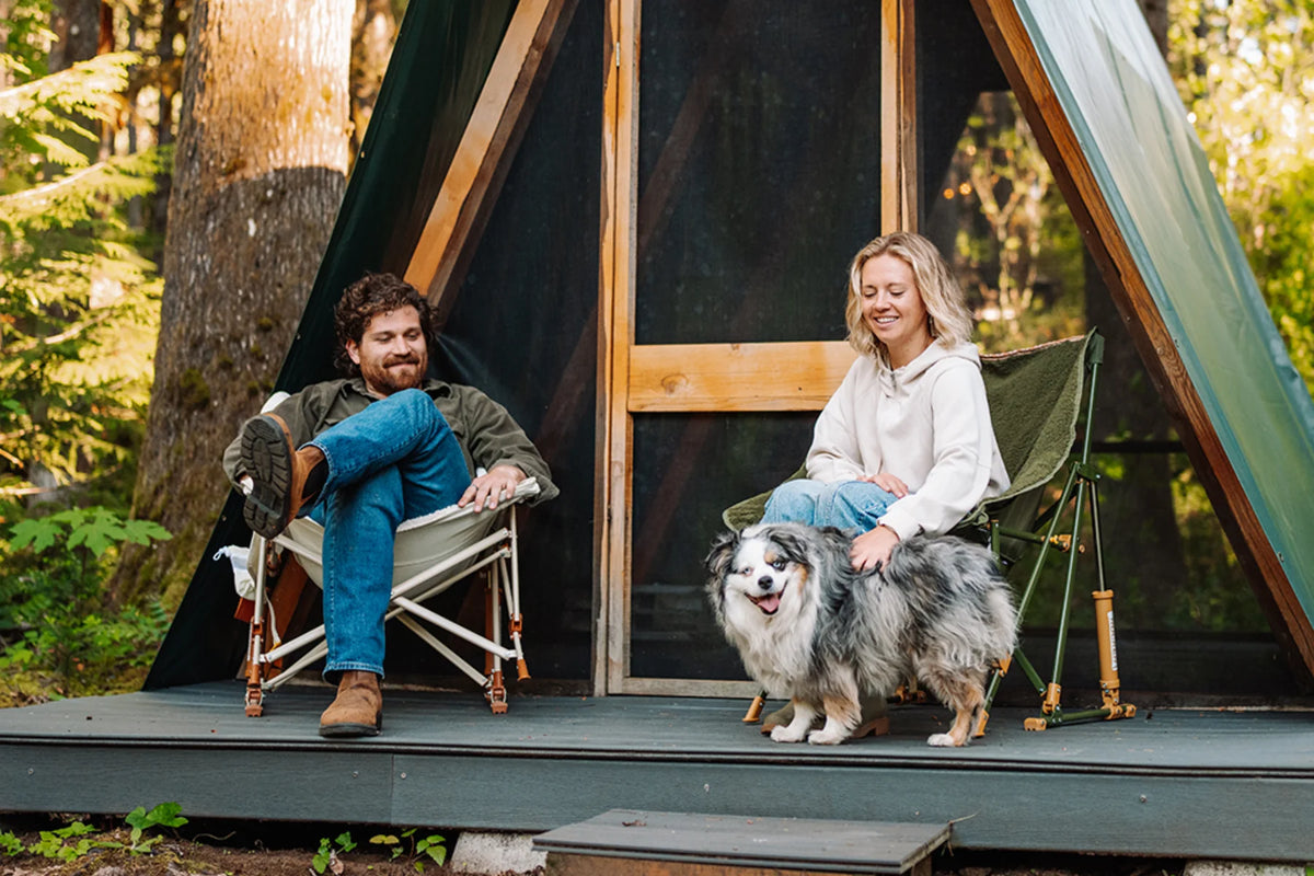 A man and woman sit in natural and olive Fleece Rocker camp chairs on a wooden platform outside an A-frame cabin, petting a dog.