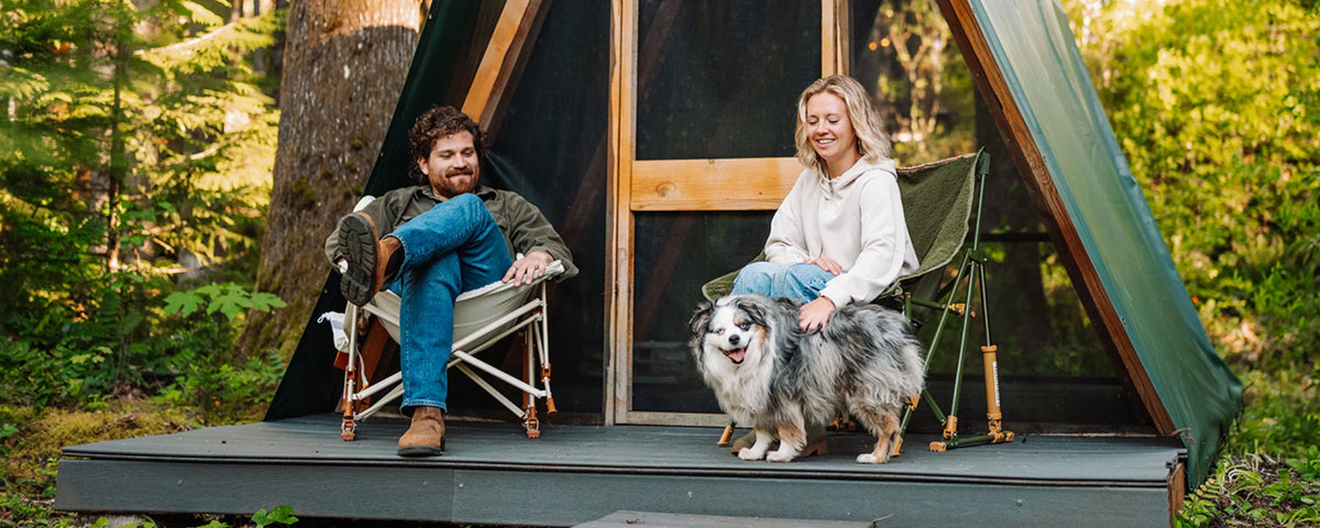 A man and woman sit in natural and olive Fleece Rocker camp chairs on a wooden platform outside an A-frame cabin, petting a dog.