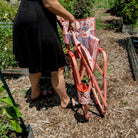 A woman collapsing a pink sundress freestyle rocker elite chair in a garden setting. 