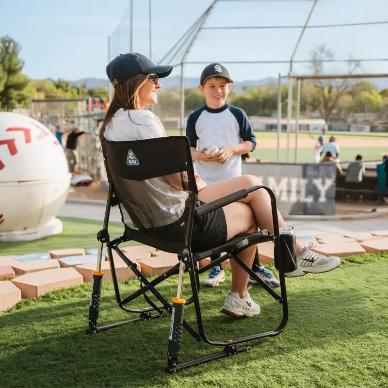 Woman relaxing in a black Freestyle Rocker Elite chair at a baseball field, chatting with a young boy during a game.