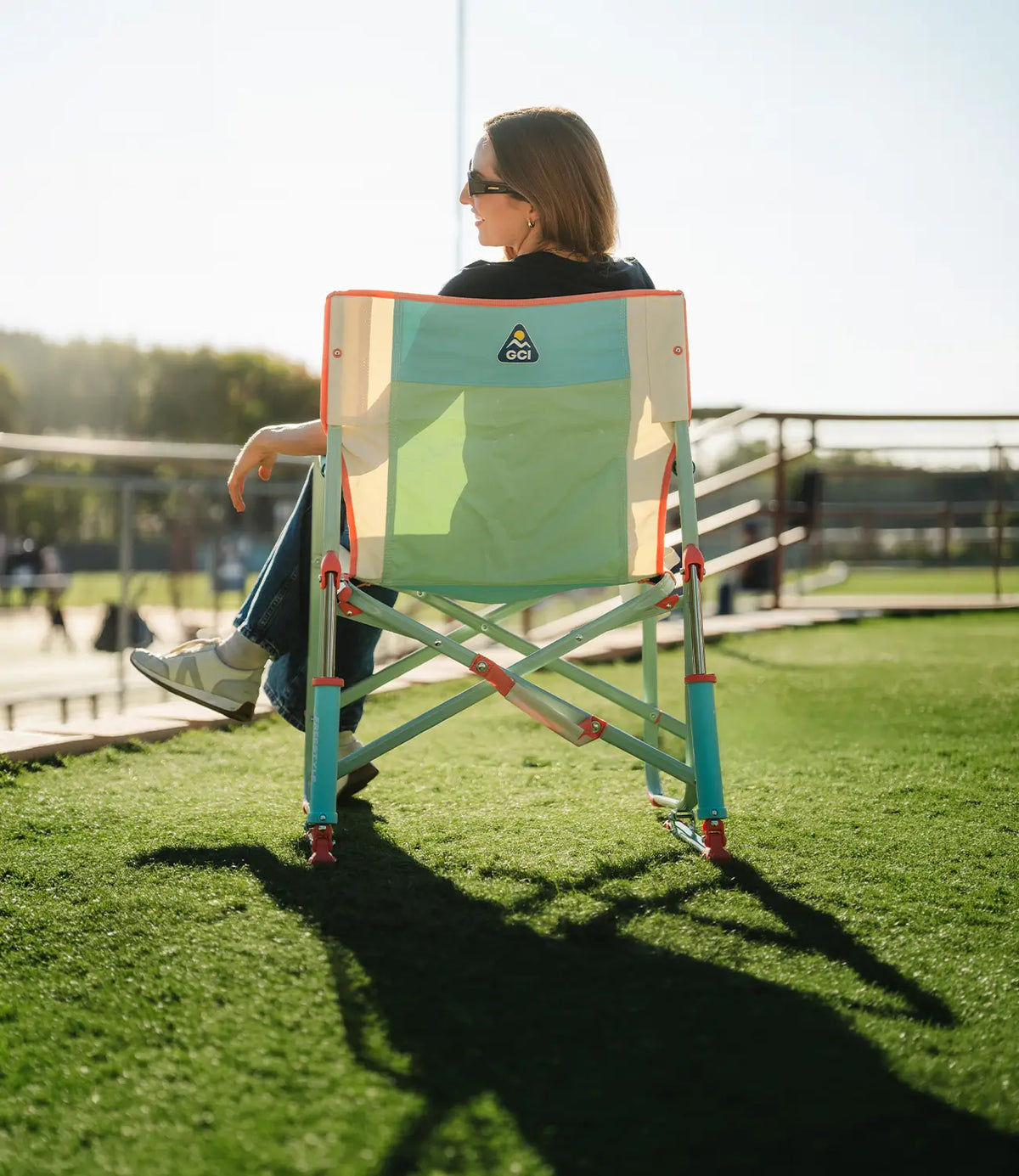 A woman sits in a Freestyle Rocker chair on green grass outdoors. The chair has a light green and teal fabric body with orange trim and a small triangular logo patch on the backrest.