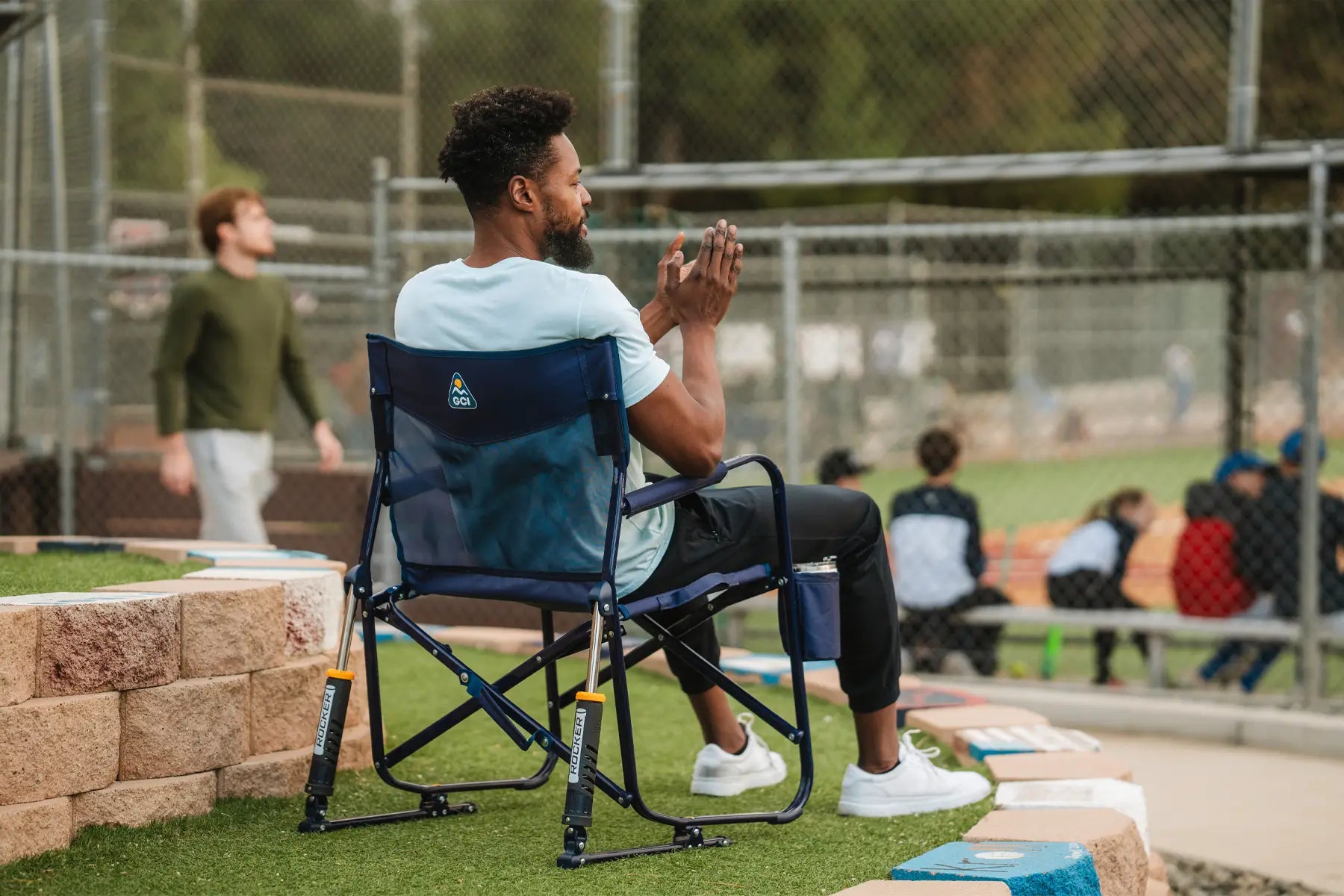 A person sitting in a blue Freestyle Rocker chair at a baseball field, watching the game comfortably.