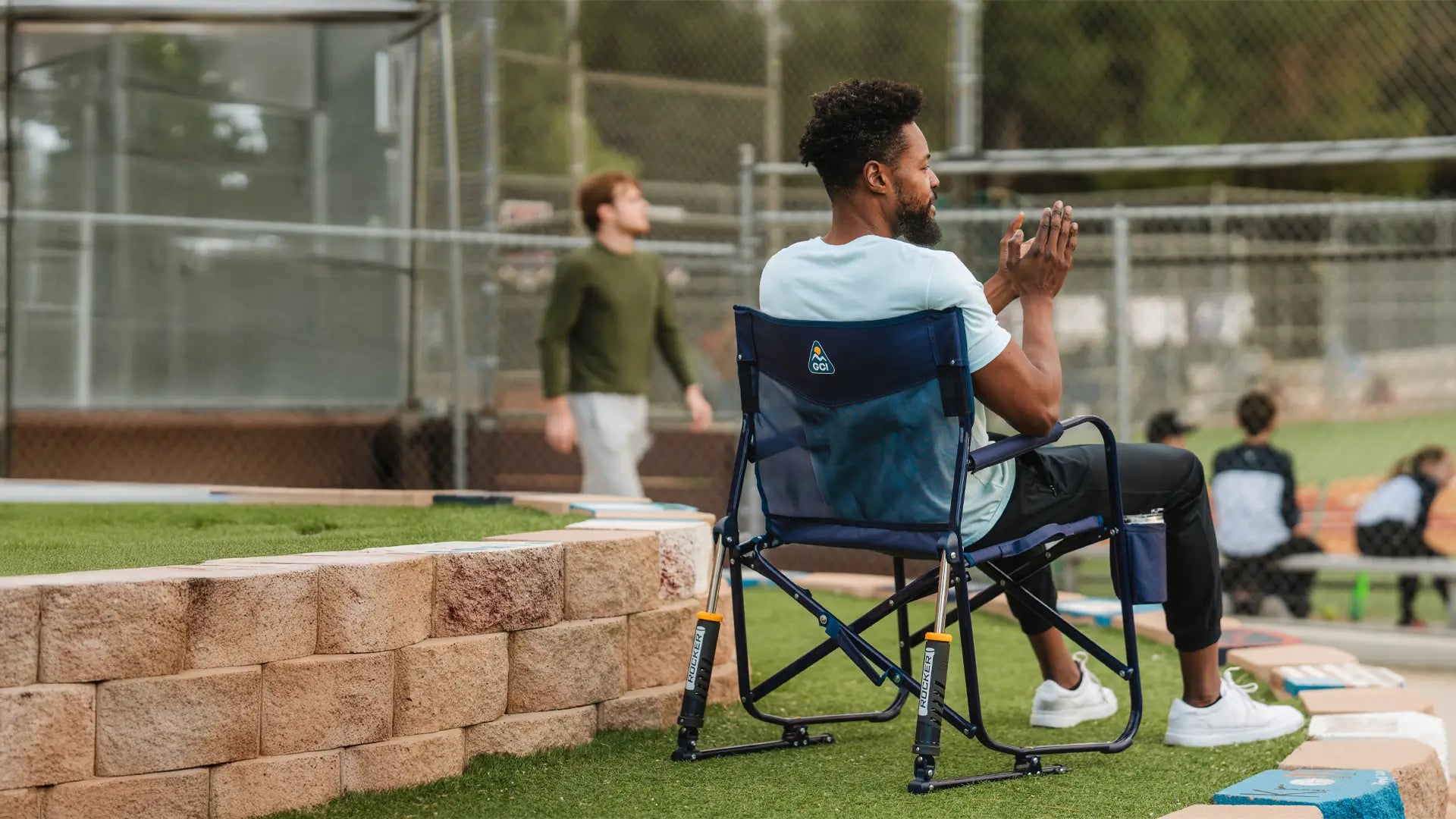 A person sitting in a blue Freestyle Rocker chair at a baseball field, watching the game comfortably.